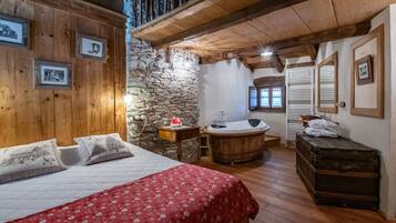 Ein gemütliches Schlafzimmer mit Holzbalkendecke, einer Steinwand, einem Bett mit roter Decke, einer Holzkiste und einem Badezimmer mit Badewanne.