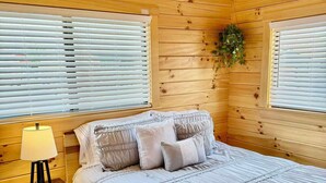 A wooden cabin bedroom with a bed, bedside table, lamp, and windows. - Walking Distance to Lakes James with Resort Amenities (Marion)