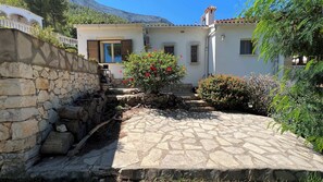 A stone-walled courtyard with a tiled floor, a small tree with red flowers, and a building with a white facade and brown shutters.