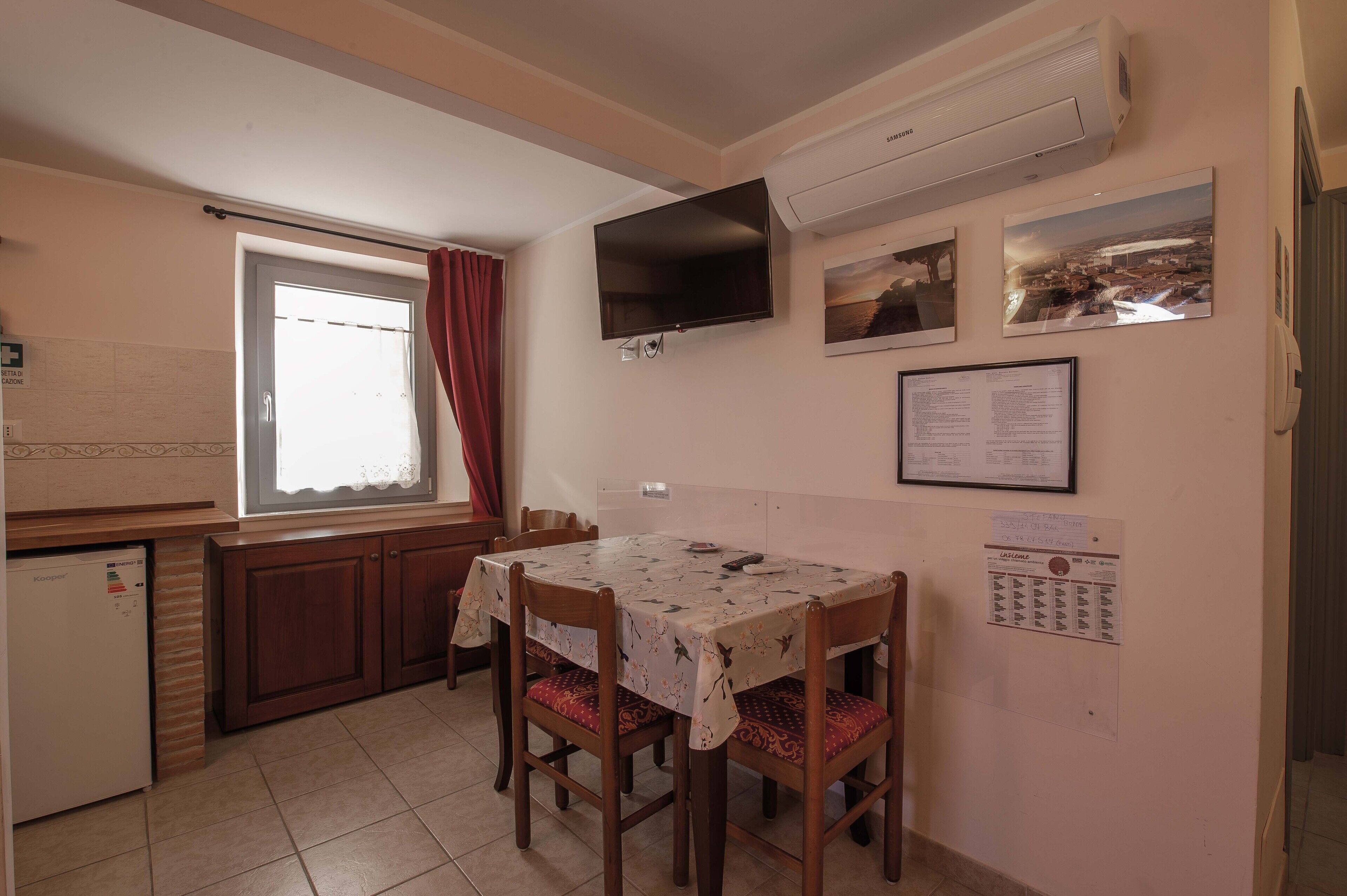 A small kitchen with a dining table, chairs, a refrigerator, and a window with curtains.