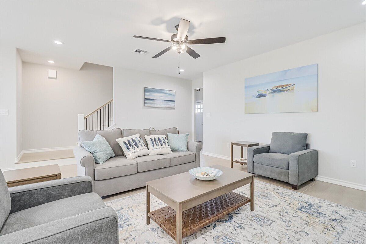 A modern living room with a grey sofa, a grey armchair, a coffee table, a ceiling fan, and a painting of a boat on the wall.