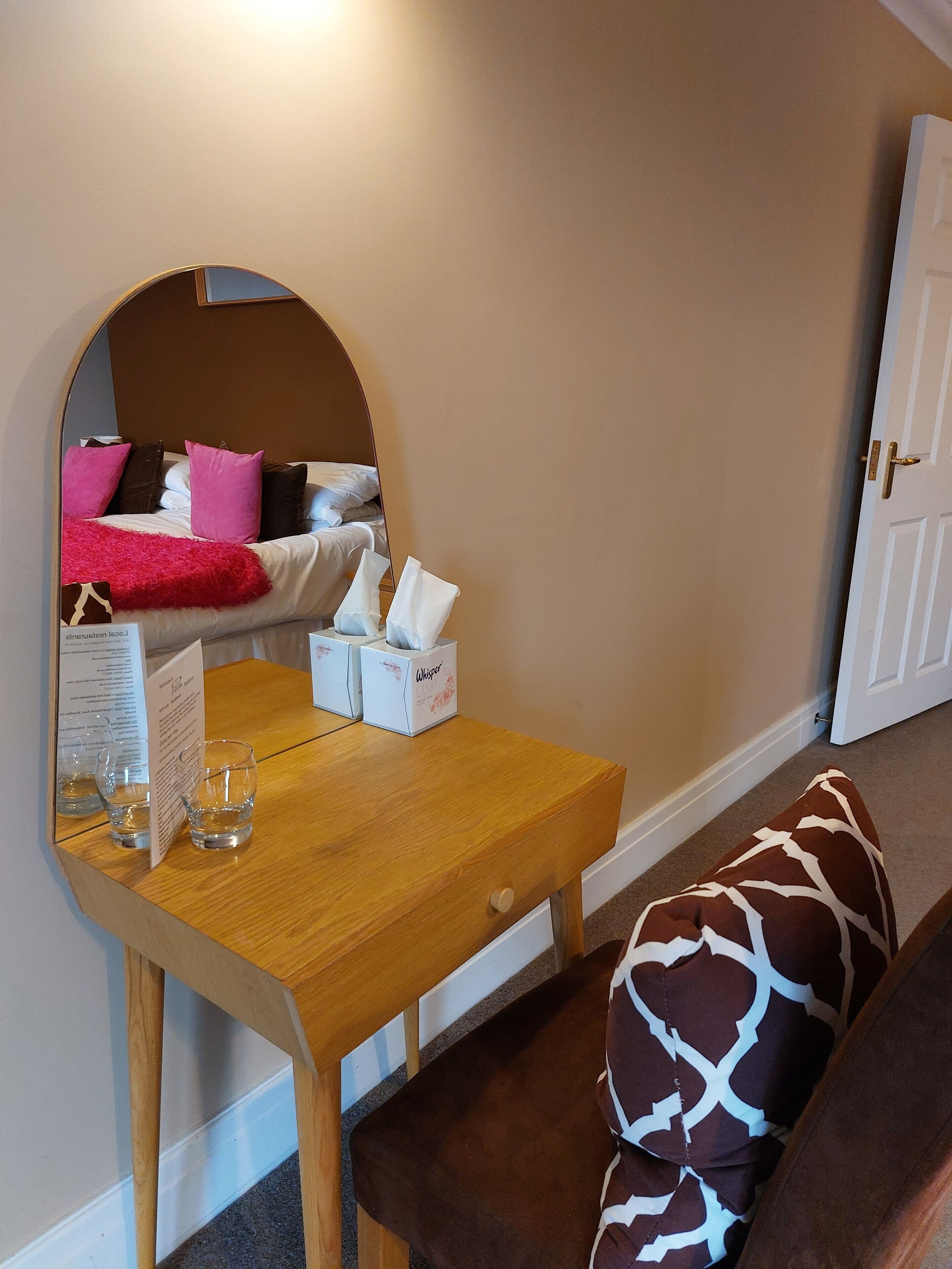 Habitación de hotel con una mesa de madera, un espejo, una cama con almohadas rosas y marrones, y una puerta.