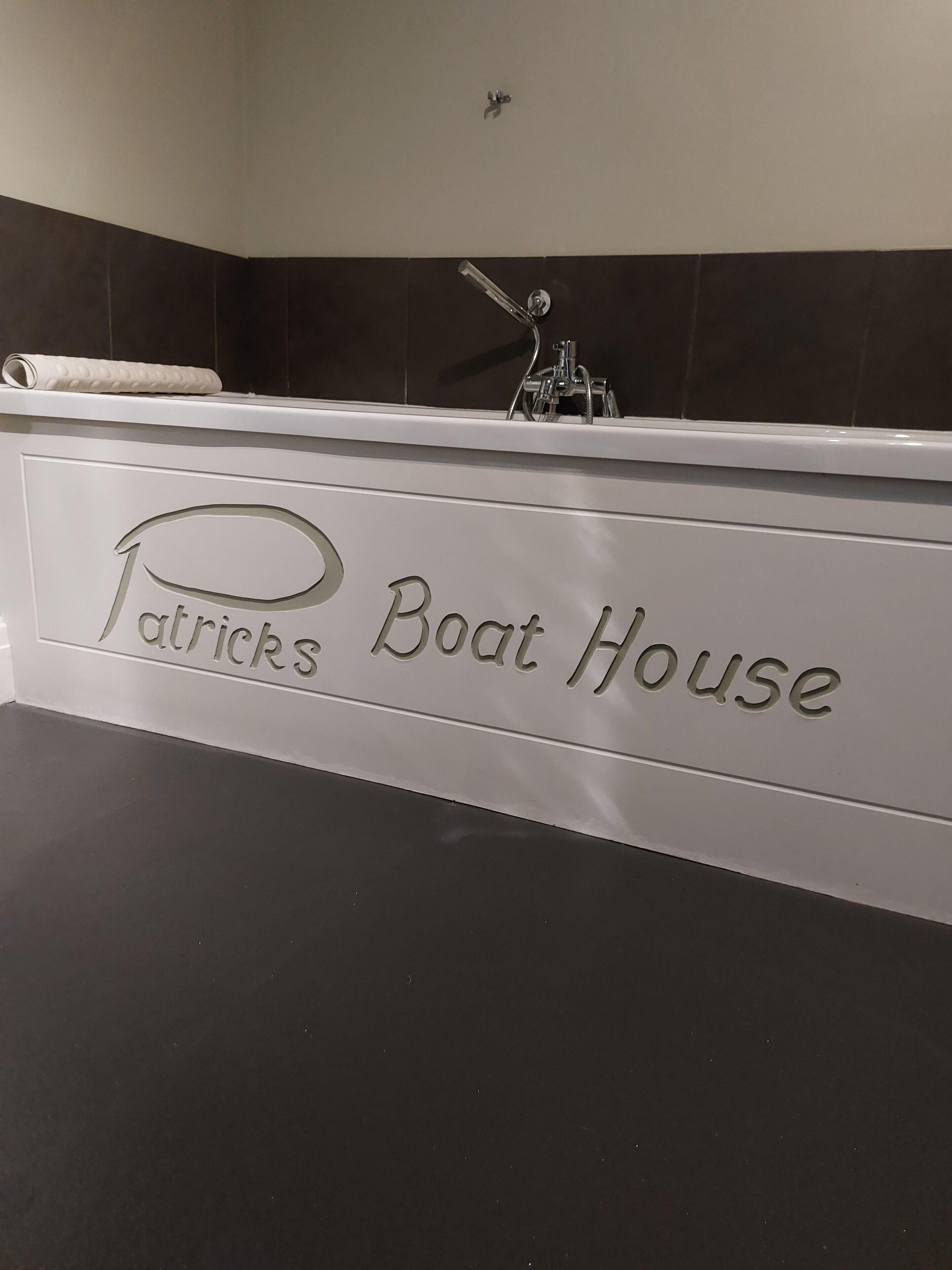 A bathroom with a bathtub, a towel, and a shower head. The wall has a dark tile pattern and a sign that reads "Patrick's Boat House".