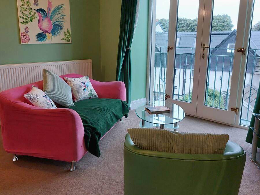 A room with a pink sofa, green armchair, and a glass table. There is a painting on the wall and a view through the glass door.