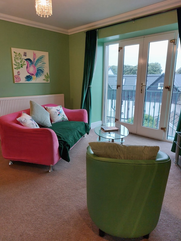 A room with a pink sofa, green armchair, and a glass table. There is a painting on the wall and a view through the glass door. - Patricks Boathouse (Swansea)