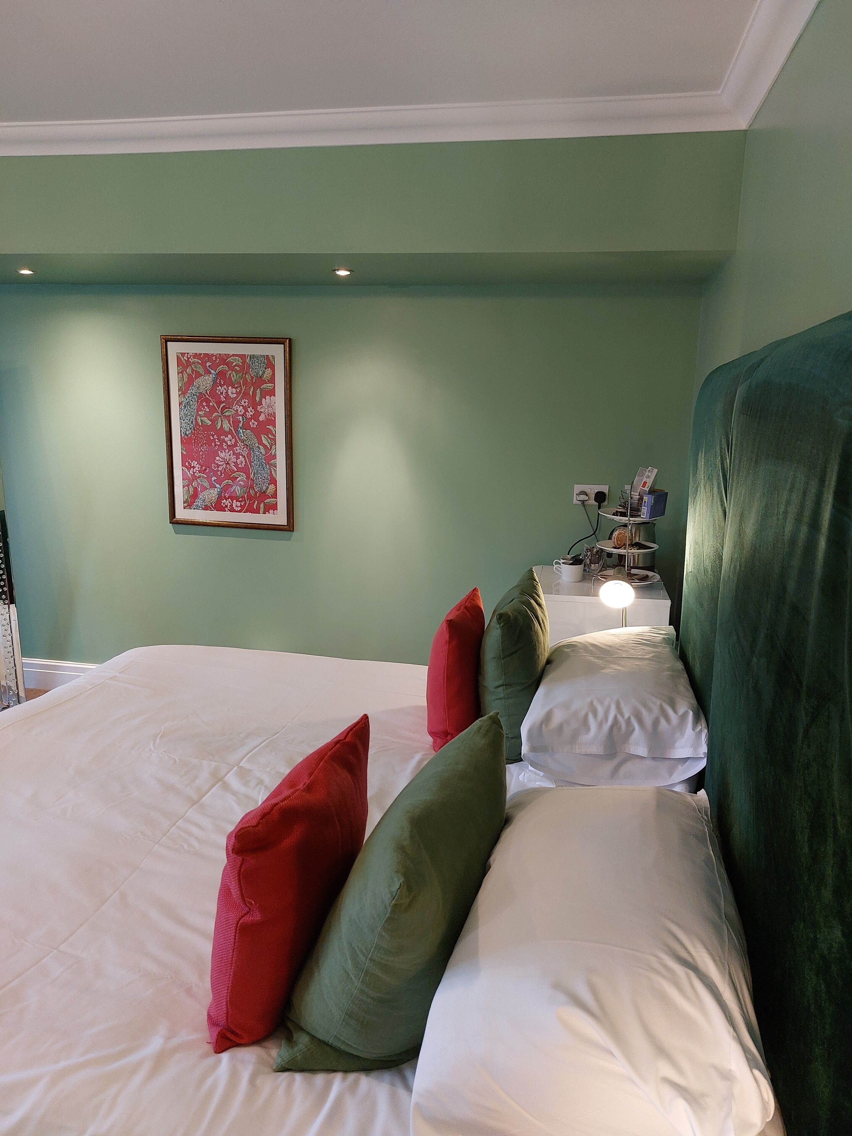 A neatly made bed with white and green pillows, a framed floral artwork on the wall, and a bedside table with a lamp.