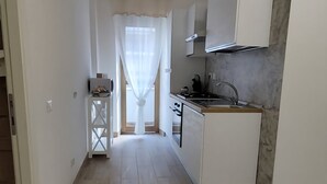 A modern kitchen with white cabinets, a stove, and a sink. There is a small island with a sink and a window with curtains. The floor is wooden, and the walls are white. - 102DreamHouse (Roma)