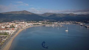 Vue aérienne d’une ville côtière avec un port de plaisance, des bâtiments le long de la côte et des montagnes en arrière-plan.