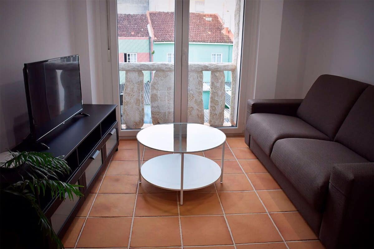Een woonkamer met een televisie, een bank, een ronde tafel en een balkon met uitzicht op gebouwen.