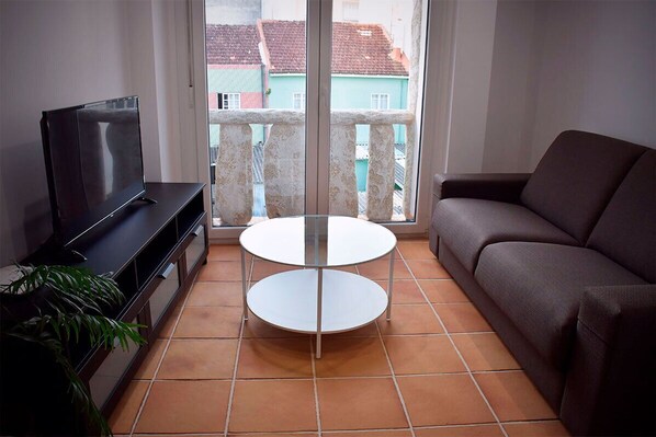 Een woonkamer met een televisie, een bank, een ronde tafel en een balkon met uitzicht op gebouwen.