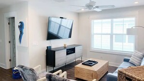 A modern living room with a flat-screen TV mounted on the wall, a ceiling fan, a sofa, a coffee table, and a large window with blinds. - Luxury 4 Bdr Beachfront in Hilton Head- DC Breeze STR Permit # 029136 (Hilton Head Island)