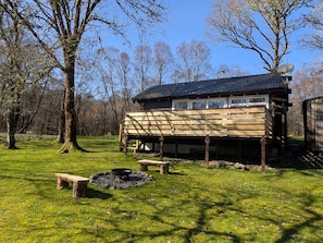 A wooden cabin with a metal roof, a fire pit, and a bench in a grassy area with trees.