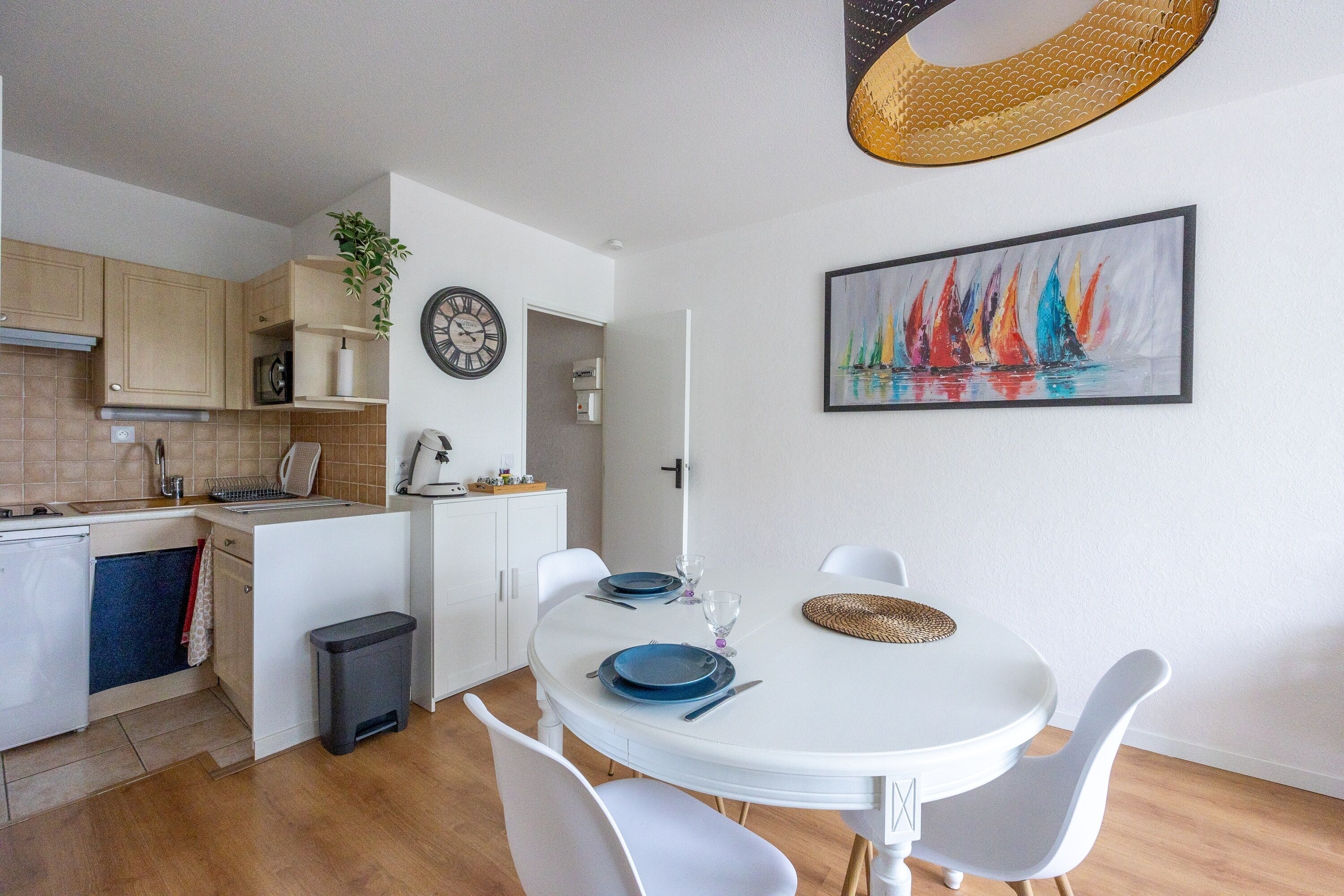 Una cocina compacta con electrodomésticos modernos, un comedor con sillas blancas y un cuadro de un velero colorido en la pared.