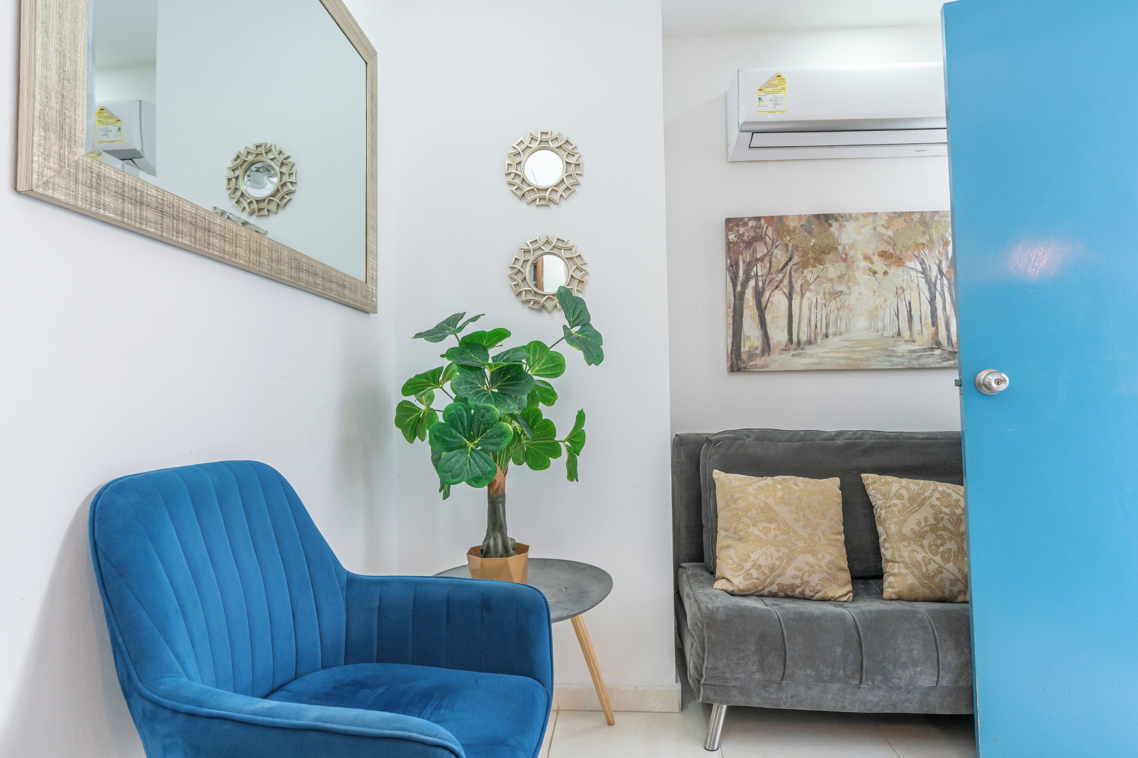 Ein Zimmer mit blauem Sessel, grauem Sofa, Topfpflanze, dekorativen Spiegeln und einem Gemälde an der Wand.