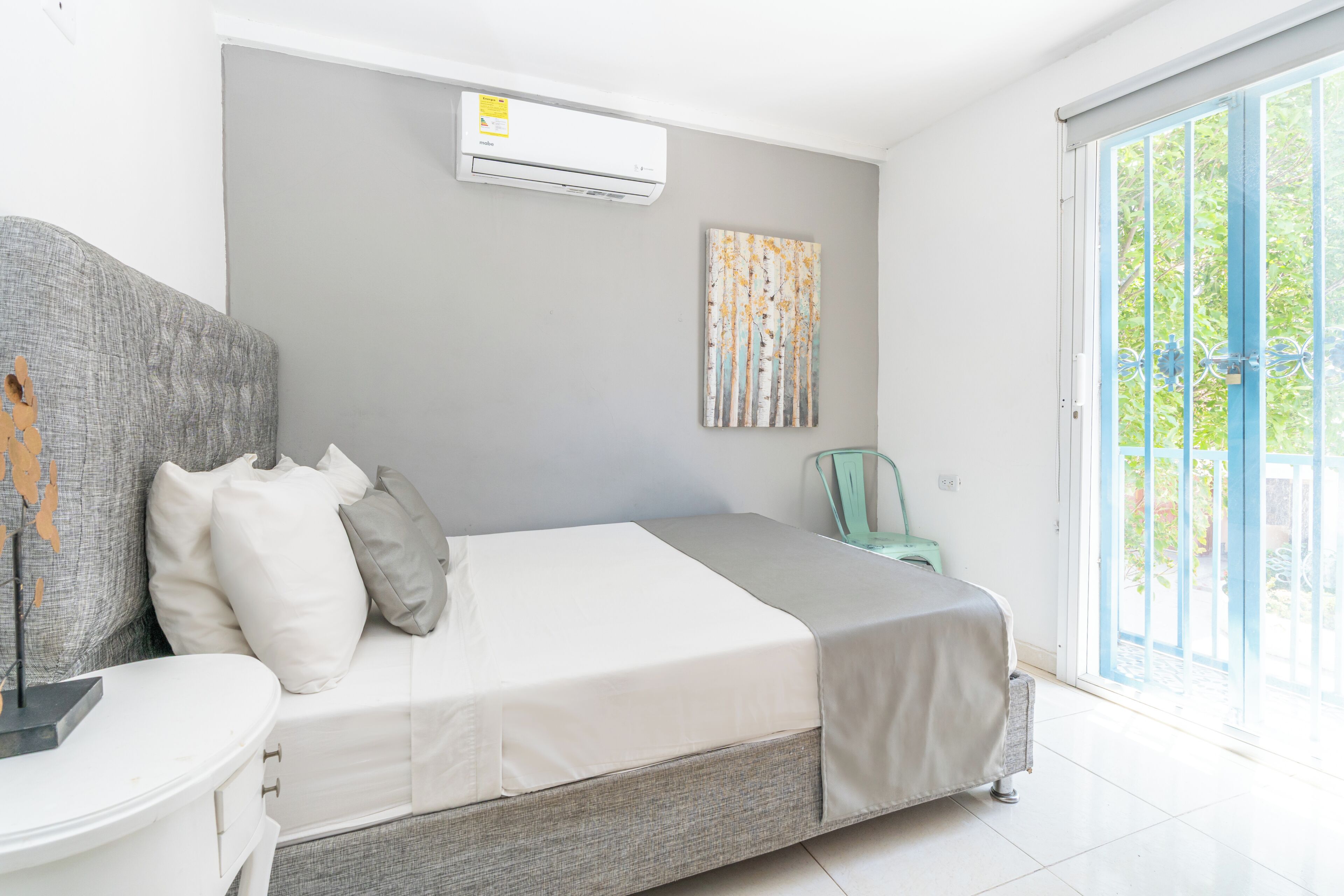 A bedroom with a bed, a bedside table, a chair, an air conditioning unit, and a view through a glass door.
