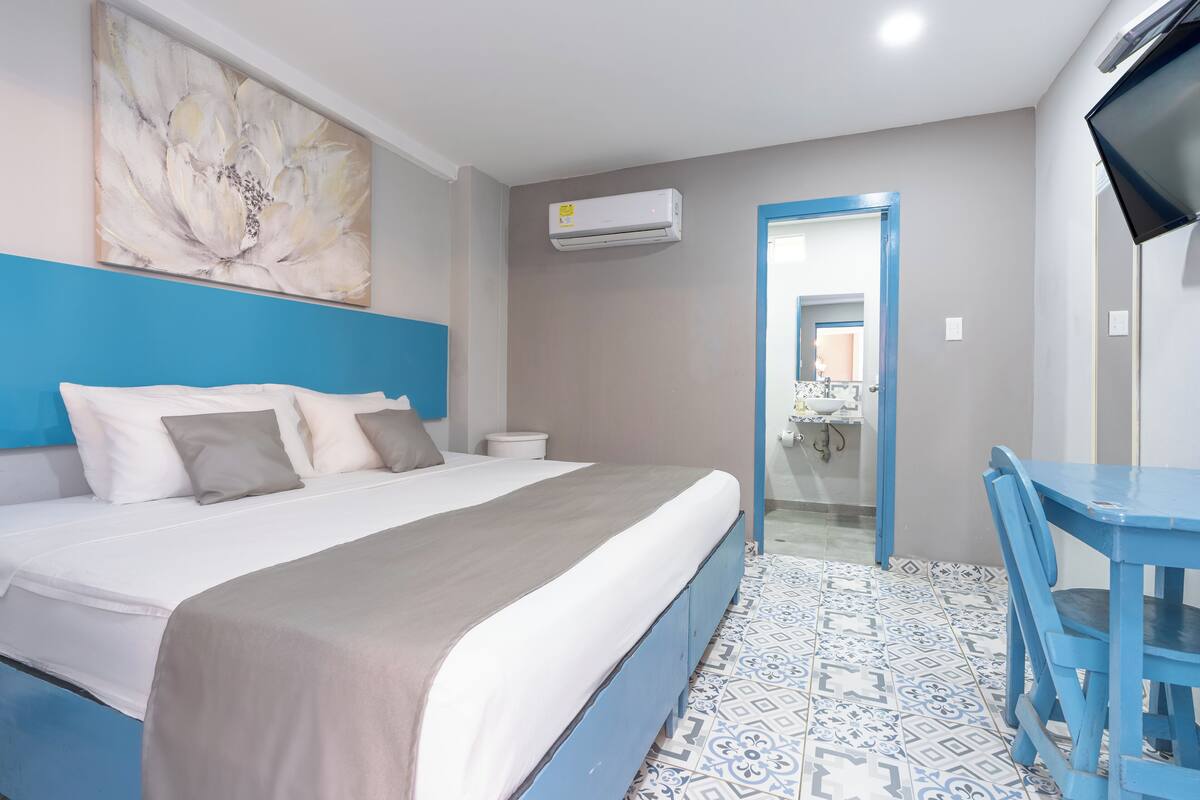 A hotel room with a bed, a desk, an air conditioning unit, and a bathroom visible through an open door.