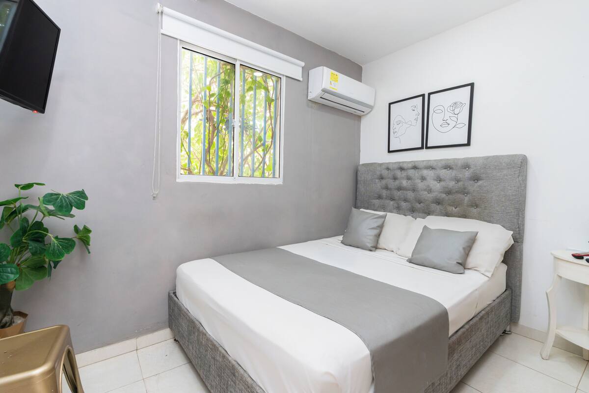 A bedroom with a bed, a window, an air conditioner, a plant, and a TV.