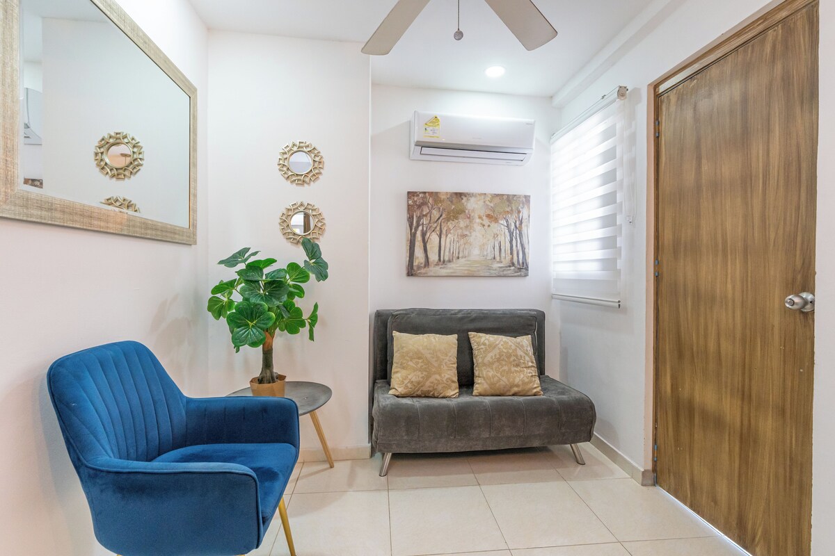A room with a blue armchair, a grey sofa with patterned cushions, a large mirror, a painting of a tree-lined path, and a wooden door.