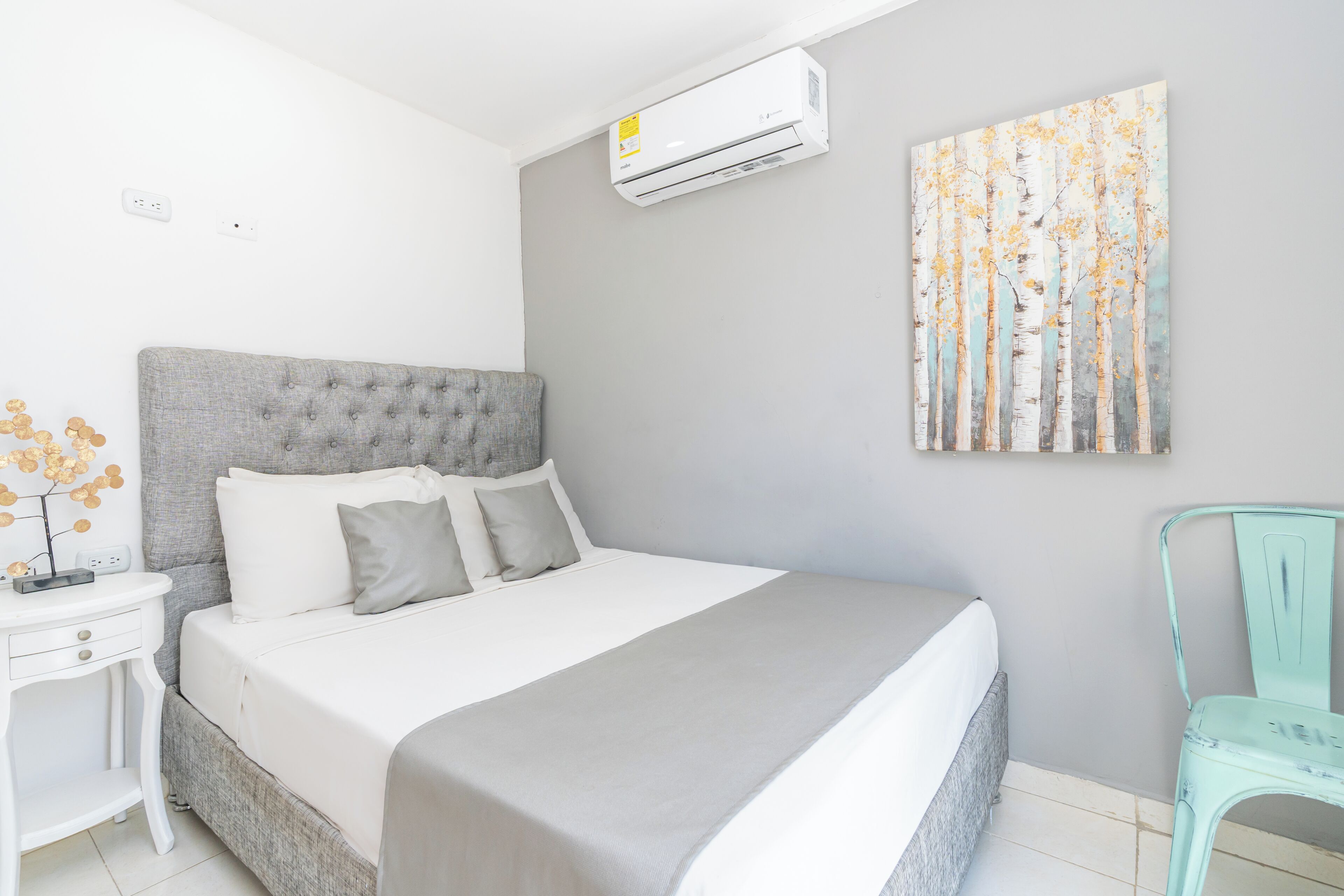 Et soveværelse med en grå polstret sengegavl, et hvidt sengetæppe, et gråt tæppe, et hvidt natbord med en lampe, og en vægmonteret aircondition.