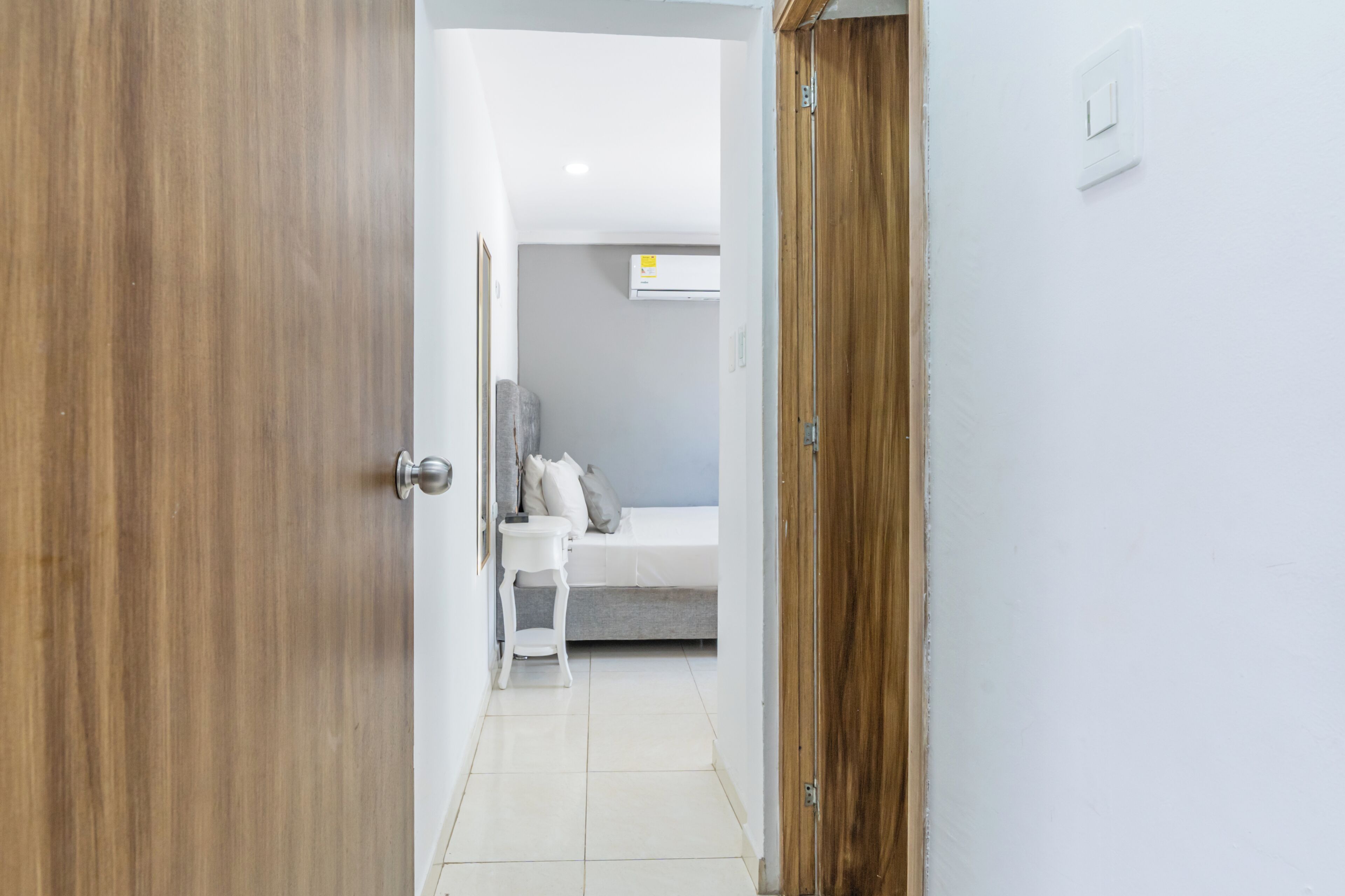 A hallway leading to a bedroom with a bed, a bedside table, and a wall-mounted air conditioner.