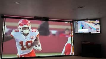 Quarto com uma grande tela de TV exibindo uma partida esportiva.