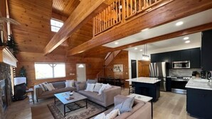 Amplia sala de estar con techo de madera, chimenea, un conjunto de sofás y una cocina equipada con electrodomésticos modernos.