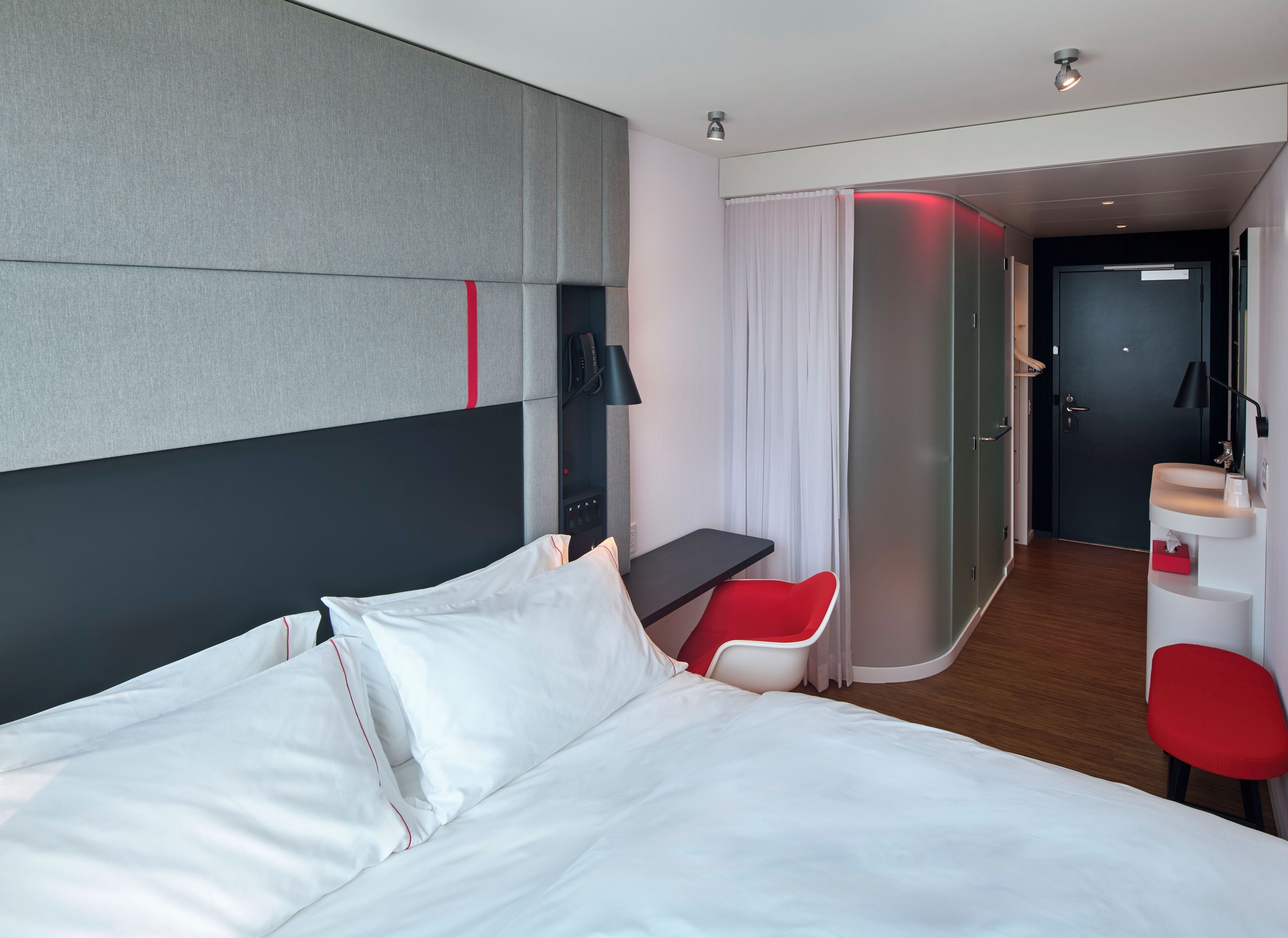A modern hotel room with a large bed, a desk, a red chair, and a bathroom visible through an open door.