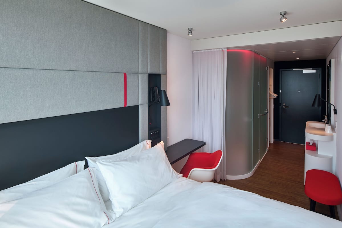 A modern hotel room with a large bed, a desk, a red chair, and a bathroom visible through an open door.