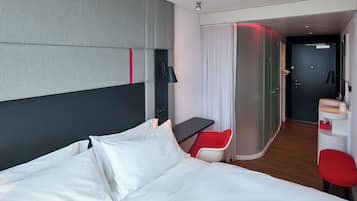 A modern hotel room with a large bed, a desk, a red chair, and a bathroom visible through an open door.
