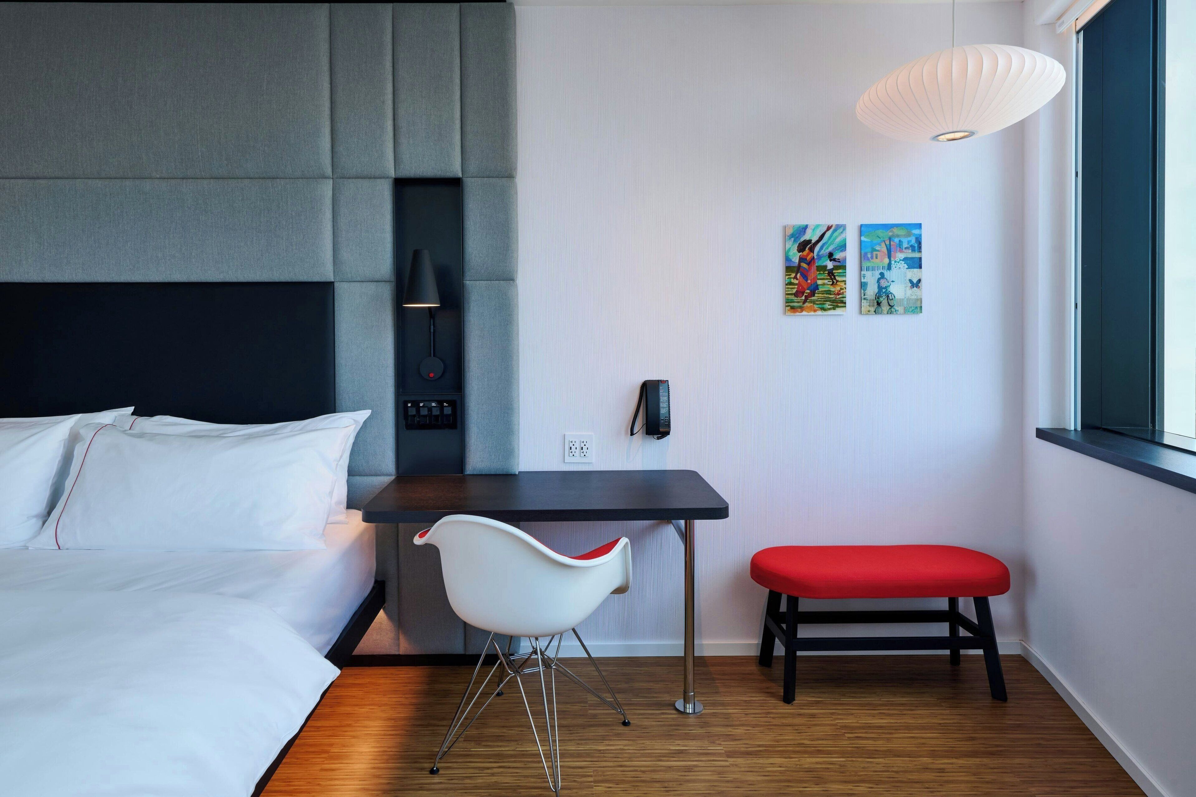 A modern hotel room with a bed, a desk, a chair, and a bench.