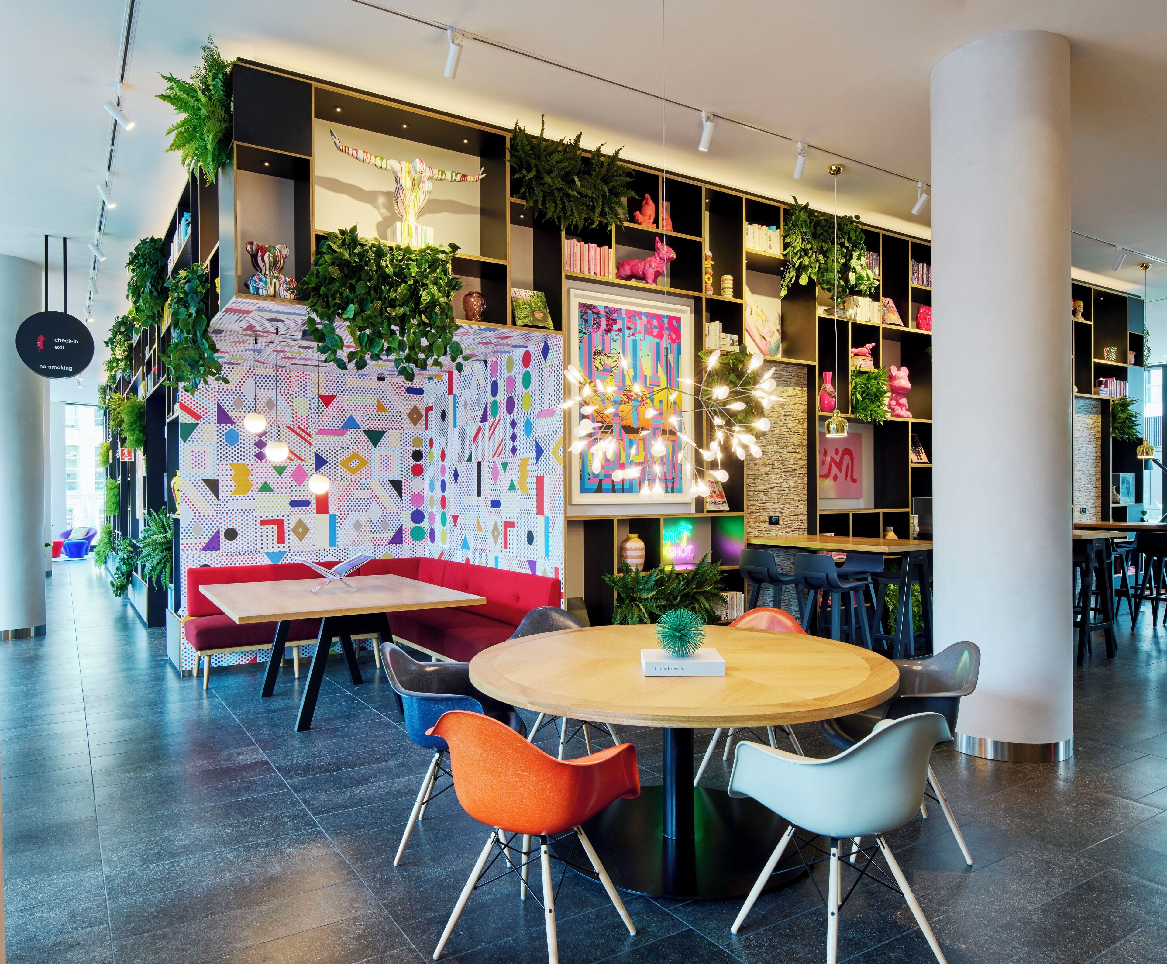 A modern lounge area with a round wooden table, colorful chairs, and a vibrant geometric wall design.