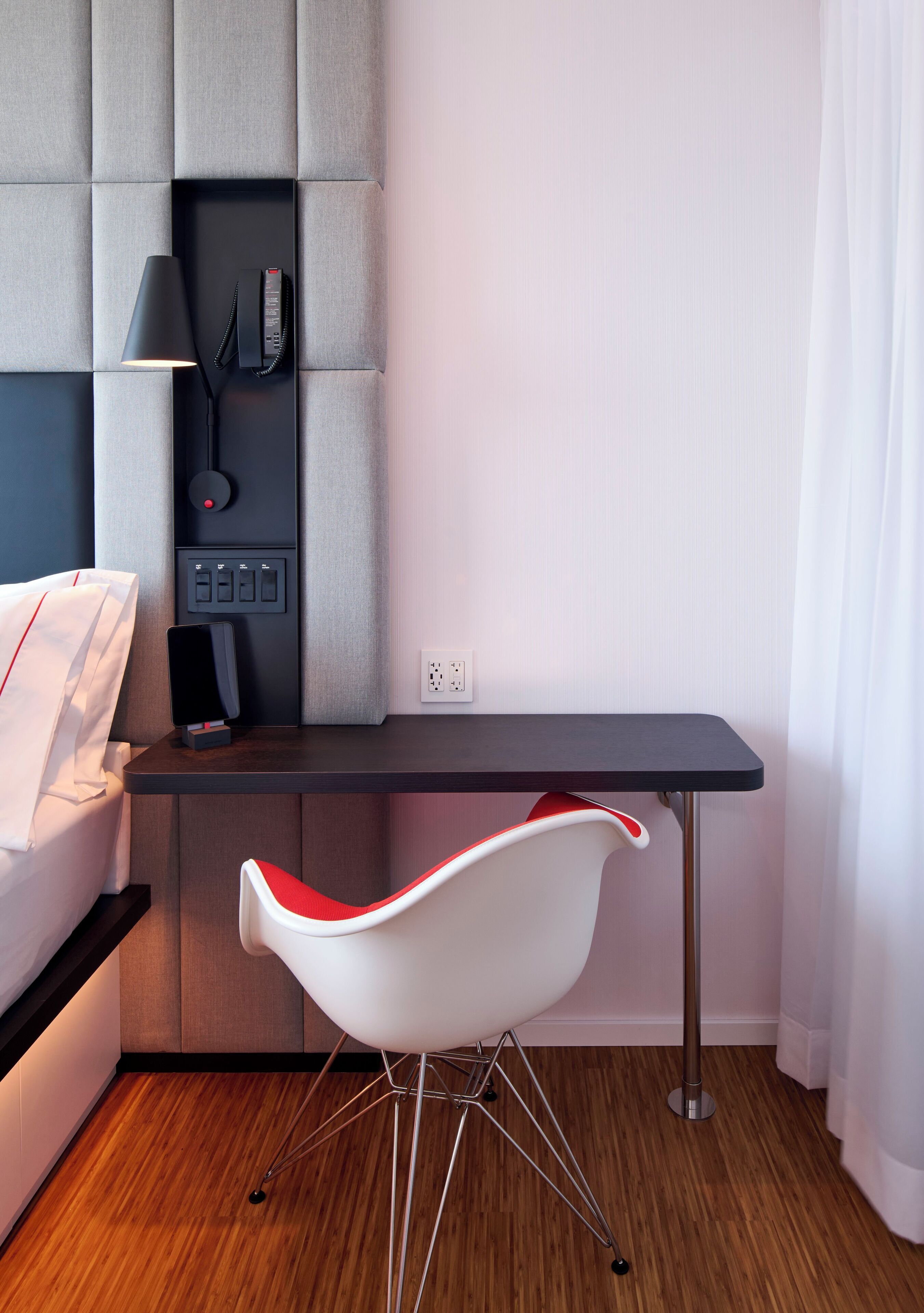 A modern hotel room with a wooden floor, a white bed, a grey padded headboard, a black desk with a lamp, a red and white chair, and a white curtain.