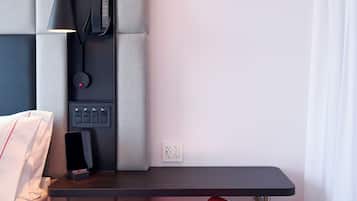 A modern hotel room with a wooden floor, a white bed, a grey padded headboard, a black desk with a lamp, a red and white chair, and a white curtain.