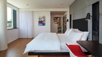A modern hotel room with a large bed, a red chair, a desk, and a city view.