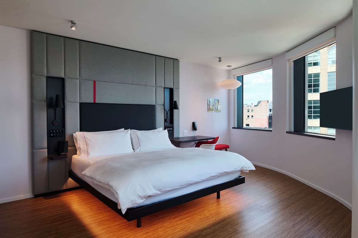 A modern hotel room with a large bed, a desk, and a view of the city through the window.