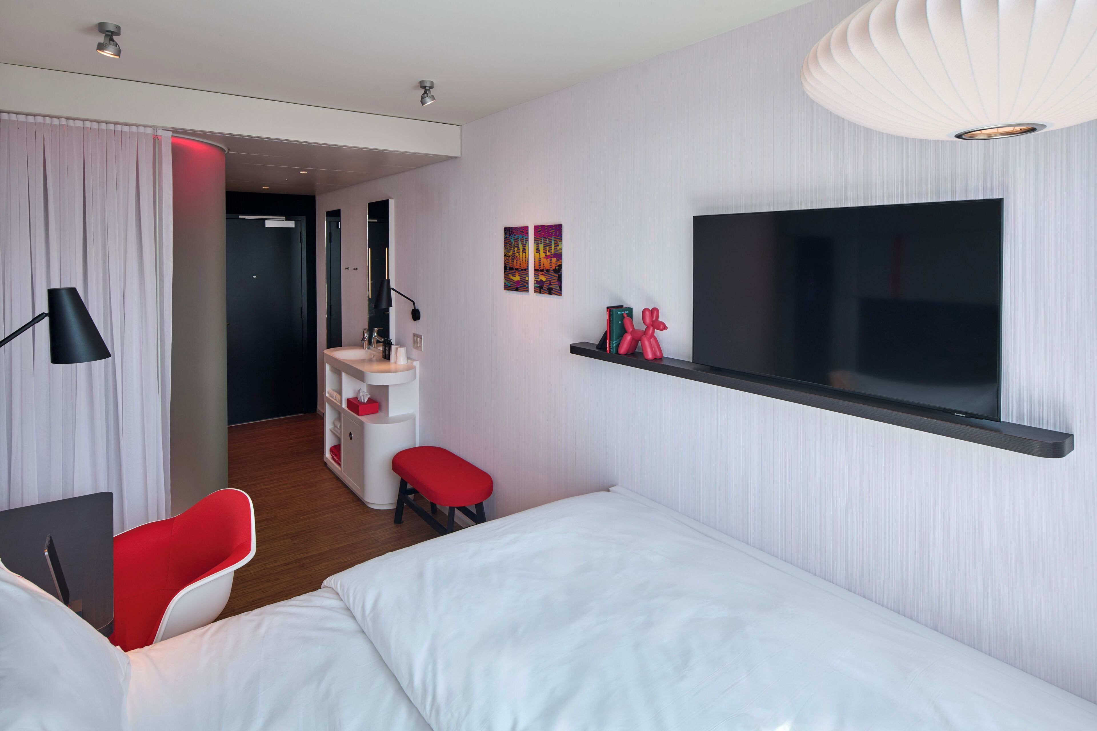 Quarto de hotel moderno com cama, escrivaninha com cadeira, TV e um pequeno espaço de estar.