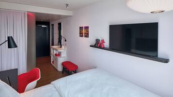 A modern hotel room with a bed, a desk with a chair, a TV, and a small seating area.