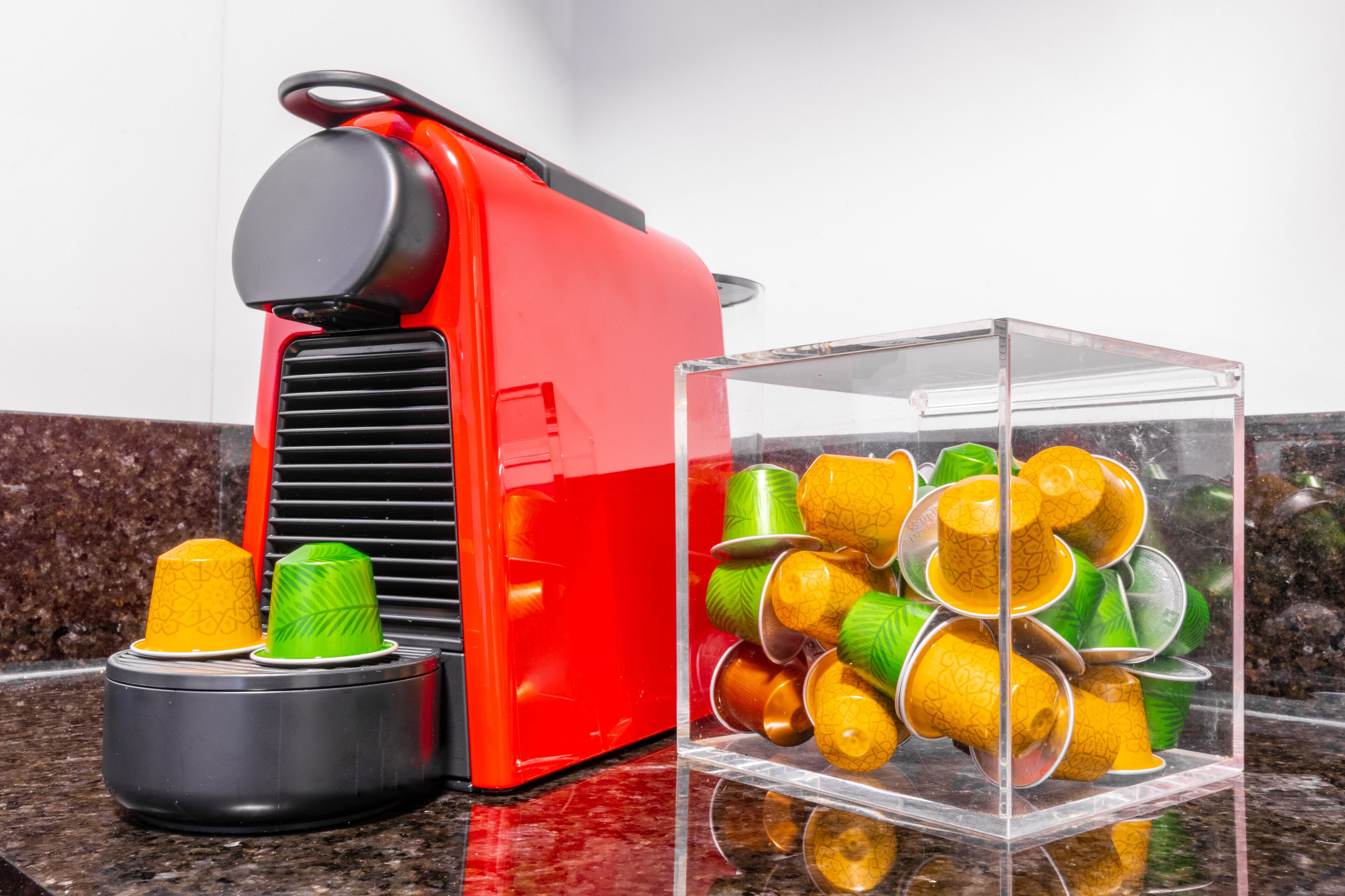 A red coffee machine and a clear glass container with colorful cups on a marble countertop.