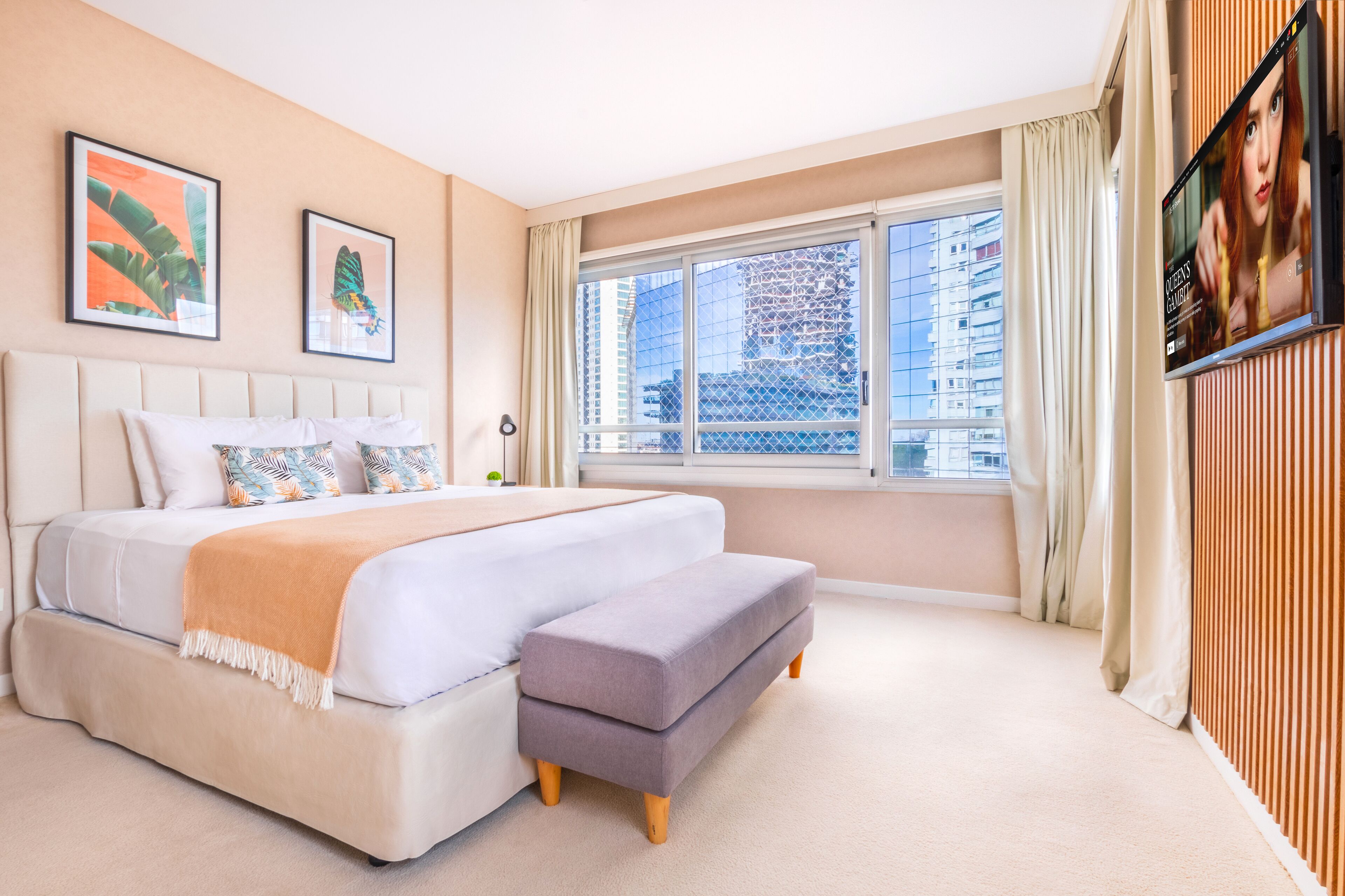 Quarto com uma cama grande, um banco, uma televisão e uma janela com cortinas.