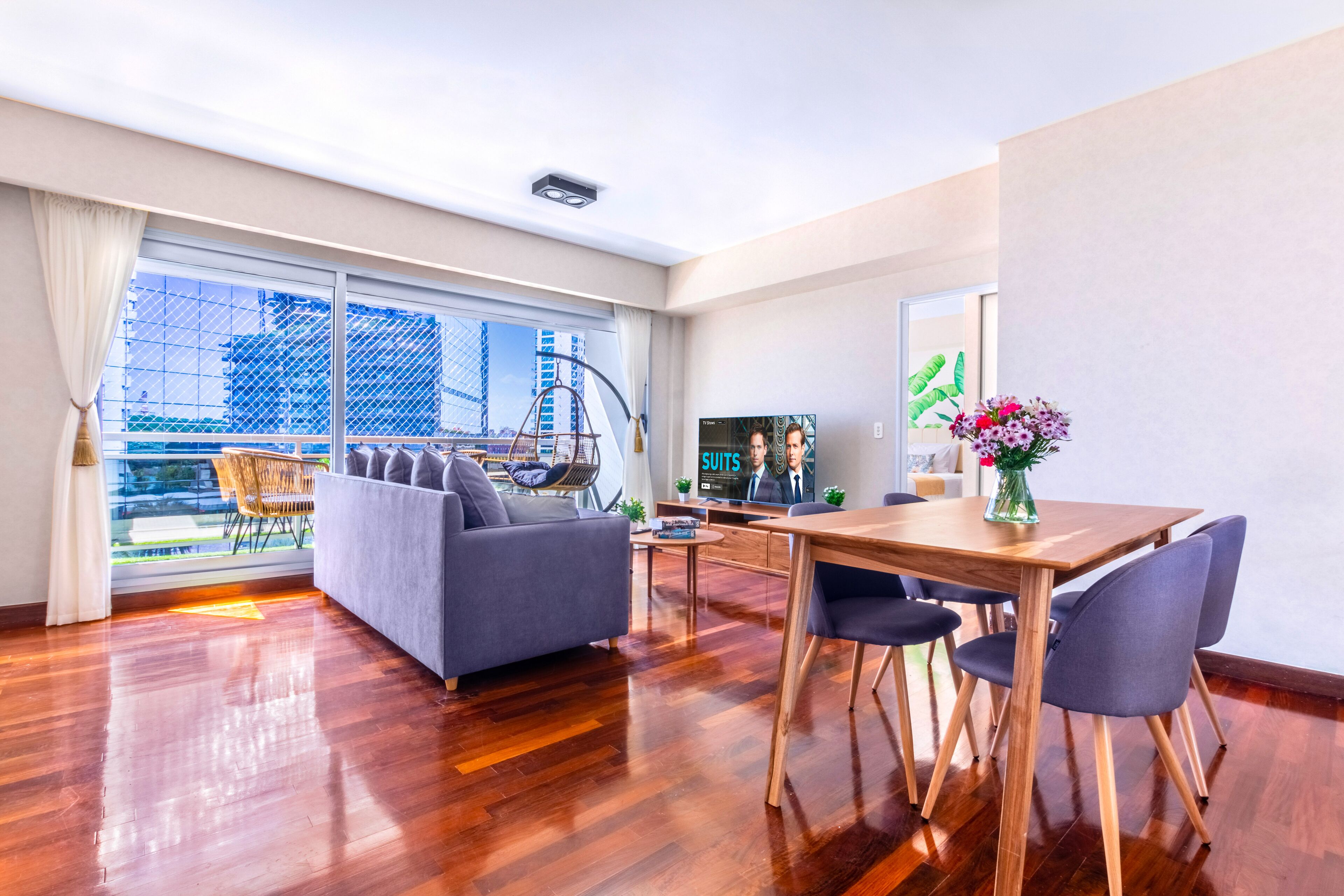 A modern hotel room with a balcony, a sofa, a dining table, and a TV.