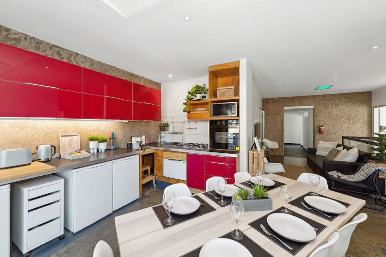 A modern kitchen with red cabinets, a dining area with a table set for four, and a living area with a sofa and a TV.