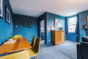 A room with a blue wall, a wooden dining table with yellow chairs, a cabinet, and a window with curtains. - ZooBlu: Chester 1-Bed (Cheshire West and Chester)