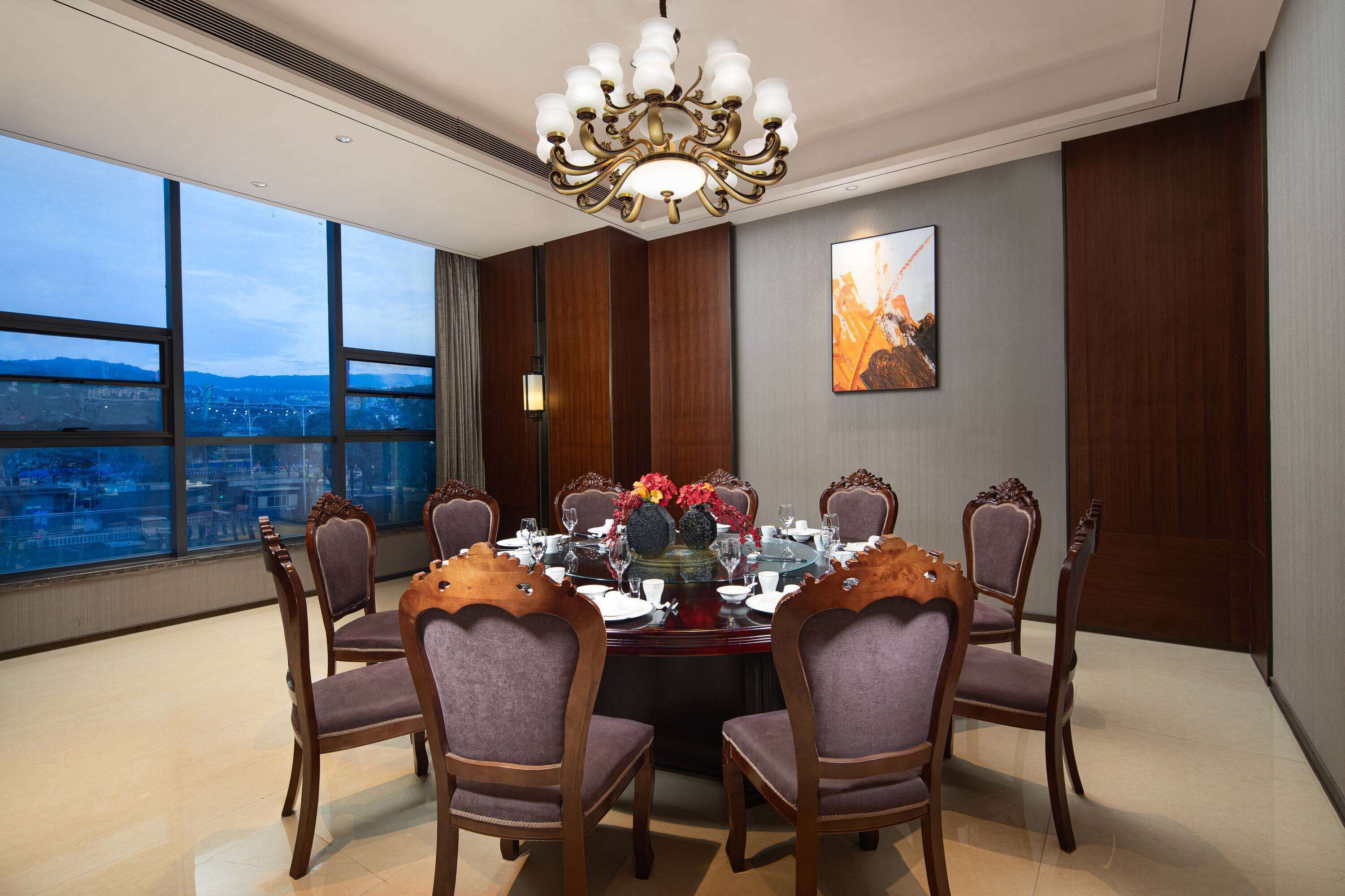 Comedor con una mesa redonda dispuesta para seis personas, rodeada de sillas de madera, un candelabro y vistas a la ciudad a través de amplios ventanales.