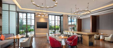 A modern hotel lobby with a herringbone wood floor, red and white seating, a large TV screen, and large windows offering a view of the outdoors.