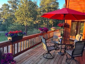 Relax on the deck with a round table and two chairs, surrounded by lush greenery and a red umbrella for shade.