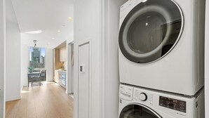 A laundry room with stacked washers and dryers, a view of the city through a window, and a kitchen area visible in the background. - Level Spacious one bedroom suite with king bed, walk-in closet, full kitchen. (Chicago)
