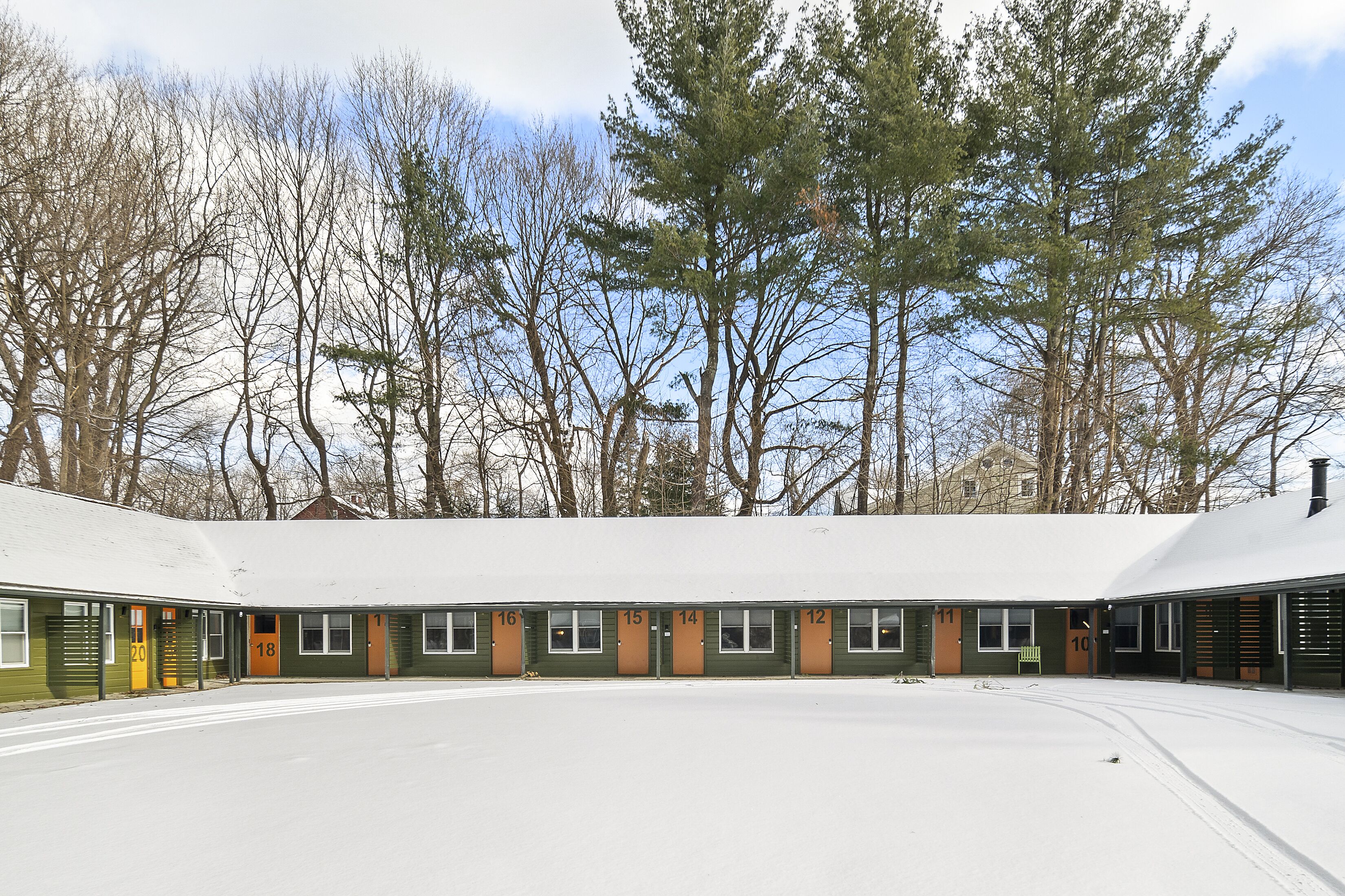 A motel with numbered rooms, a covered parking area, and a snowy landscape with trees in the background.