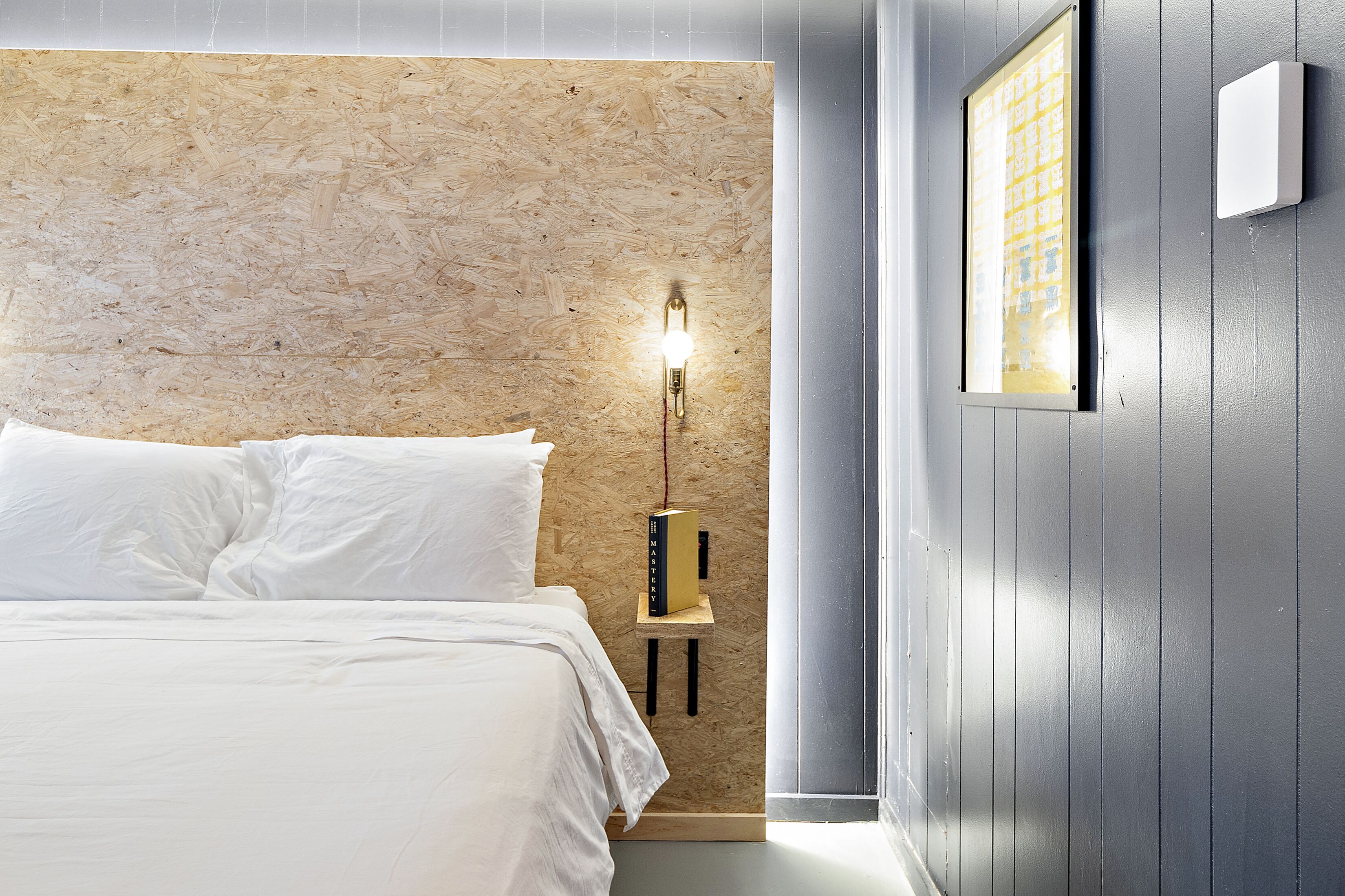 A bedroom with a wooden wall, a bed with white linens, a bedside table with a lamp, and a framed picture on the wall.