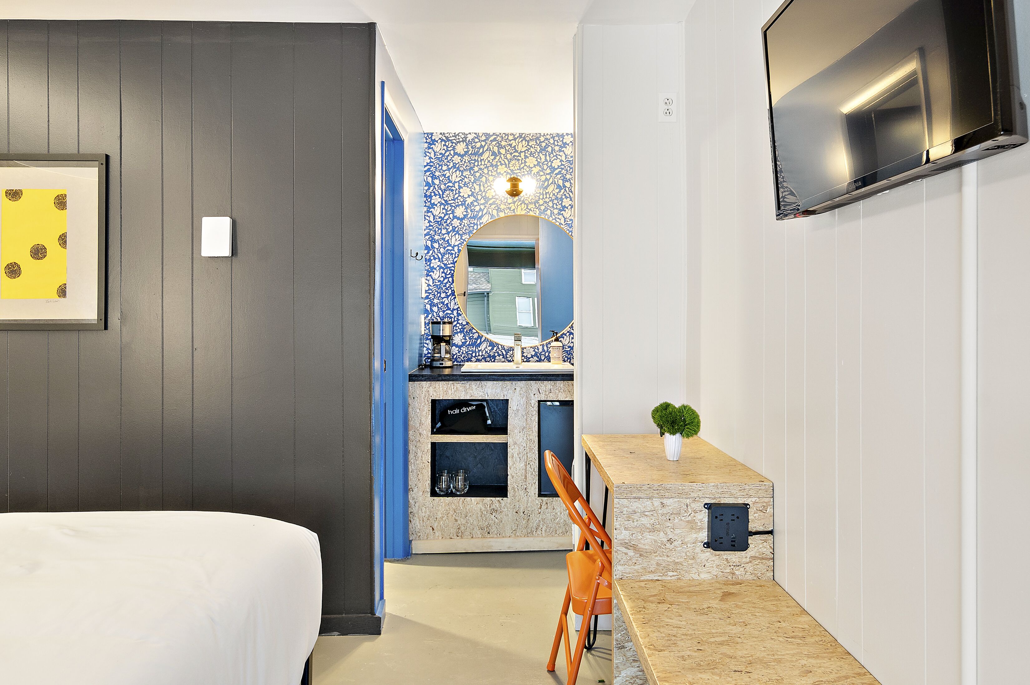 Ein Hotelzimmer mit einem Bett, einem Schreibtisch mit Stuhl, einem an der Wand befestigten Fernseher und einem Badezimmer mit Spiegel und Waschbecken.