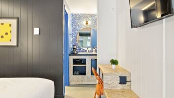 Ein Hotelzimmer mit einem Bett, einem Schreibtisch mit Stuhl, einem an der Wand befestigten Fernseher und einem Badezimmer mit Spiegel und Waschbecken.
